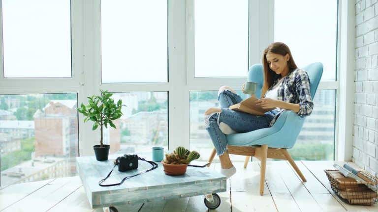 Young attractive woman read book and drink coffee sitting on balcony in modern loft apartment